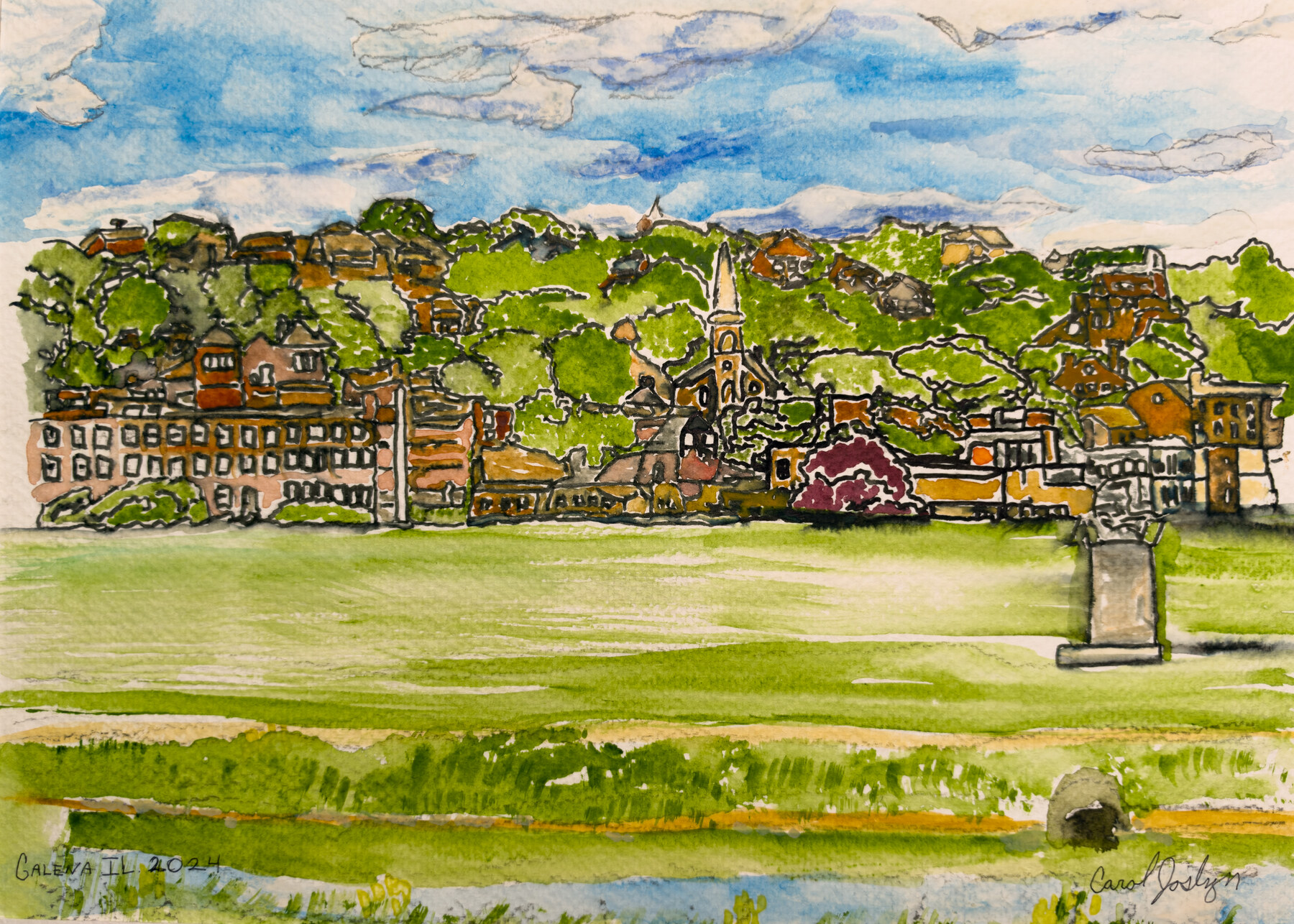 Buildings and a church in Galena sit across a broad green lawn beneath wooded hills and a blue sky.